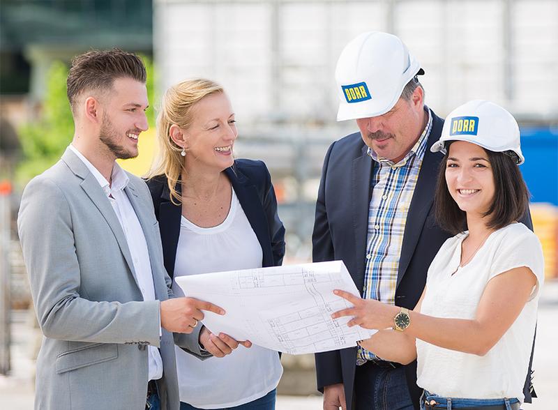 Foto: vier lächelnde Menschen in legeren Business-Outfits, die gemeinsam einen Bauplan ansehen; zwei davon mit PORR Helmen